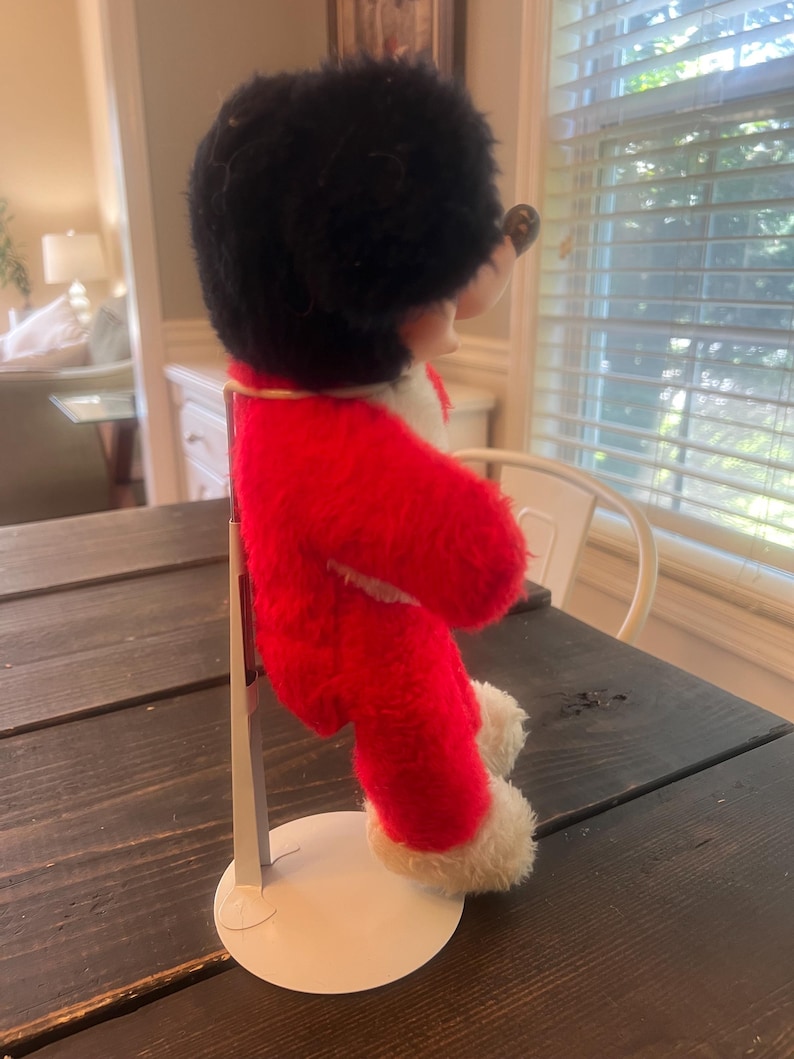 May include: A vintage plush toy of a mouse character, dressed in a red coat with white trim and a black furry hat. The toy is mounted on a white metal stand, showcasing its side profile. The toy's features include a round black nose and small ears.