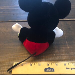 May include: Black and red plush Mickey Mouse toy with a long black tail. The toy is sitting on a wooden surface with a ruler measuring 7 inches.