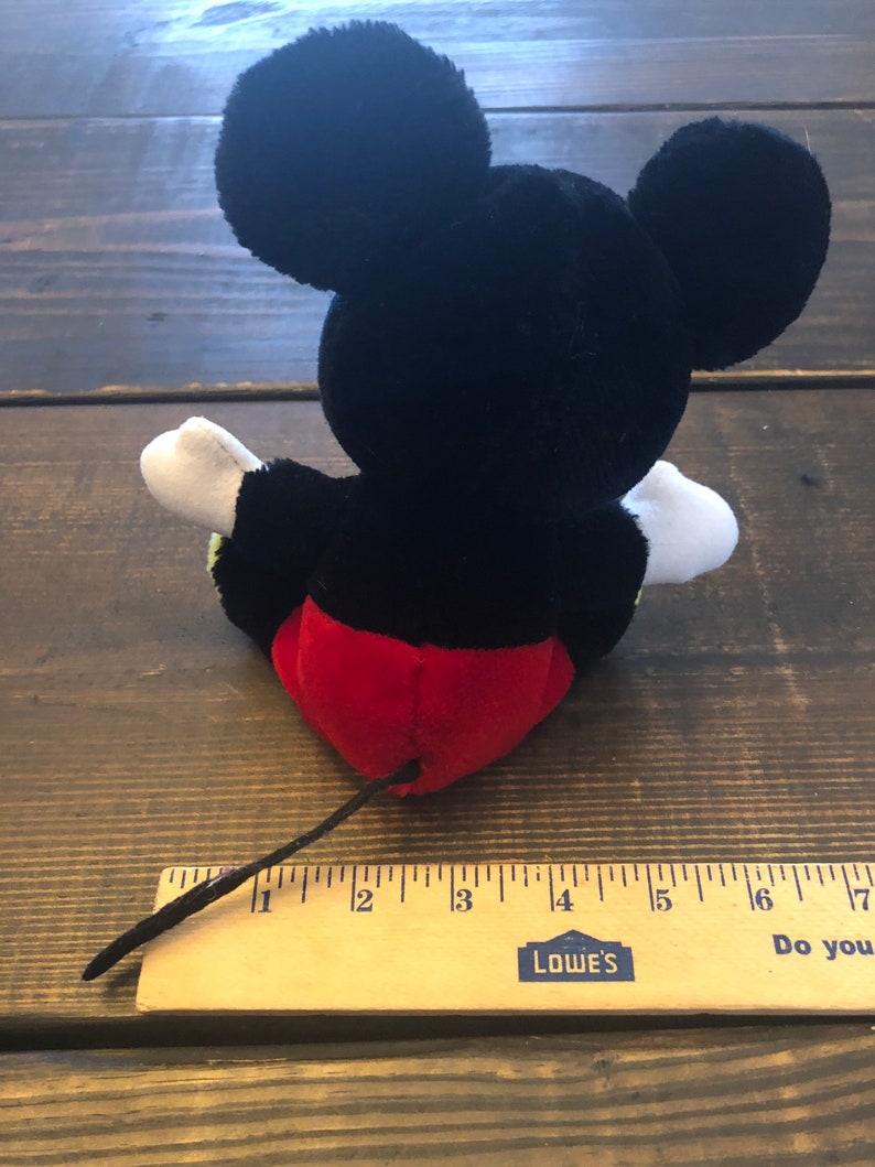 May include: Black and red plush Mickey Mouse toy with a long black tail. The toy is sitting on a wooden surface with a ruler measuring 7 inches.