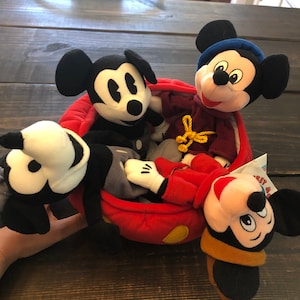 Peut inclure: Quatre peluches Mickey Mouse dans une voiture rouge en peluche. Les peluches Mickey Mouse sont toutes de designs diff&eacute;rents, dont un Mickey Mouse classique noir et blanc, un Mickey Mouse portant un chapeau bleu et un Mickey Mouse portant un chapeau marron.