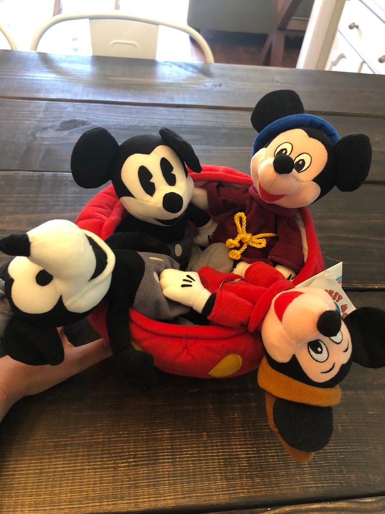 Peut inclure: Quatre peluches Mickey Mouse dans une voiture rouge en peluche. Les peluches Mickey Mouse sont toutes de designs diff&eacute;rents, dont un Mickey Mouse classique noir et blanc, un Mickey Mouse portant un chapeau bleu et un Mickey Mouse portant un chapeau marron.