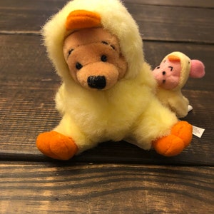 May include: A yellow plush Winnie the Pooh bear wearing a yellow duck costume. The bear is sitting with its legs crossed and has a small pink plush pig wearing a yellow duck costume sitting on its back.