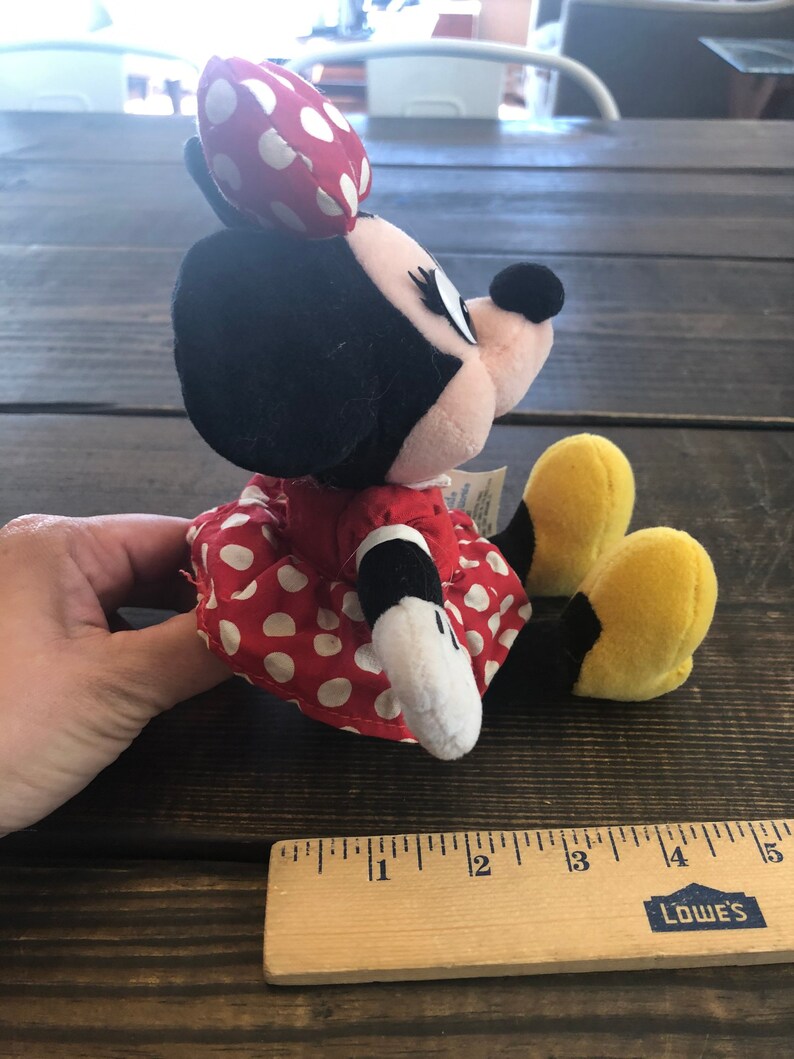 Puede incluir: Un vestido rojo con lunares blancos y una cabeza de peluche de Minnie Mouse en blanco y negro. El peluche est&aacute; sentado sobre una superficie de madera con una regla.