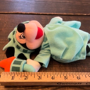May include: A plush toy of Mickey Mouse dressed as the Statue of Liberty. The toy is lying on a wooden surface with a ruler next to it. The toy is approximately 6 inches long.
