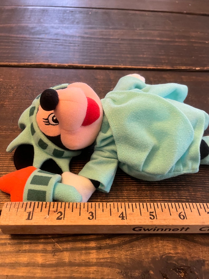 May include: A plush toy of Mickey Mouse dressed as the Statue of Liberty. The toy is lying on a wooden surface with a ruler next to it. The toy is approximately 6 inches long.