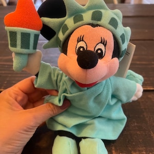 May include: A plush toy of Mickey Mouse dressed as the Statue of Liberty. The toy is holding a torch in its right hand. The toy is wearing a green dress and a crown with a green and white pattern. The toy is sitting on a wooden surface.