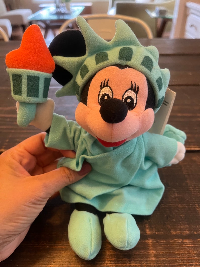 May include: A plush toy of Mickey Mouse dressed as the Statue of Liberty. The toy is holding a torch in its right hand. The toy is wearing a green dress and a crown with a green and white pattern. The toy is sitting on a wooden surface.