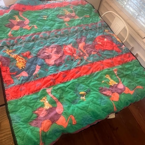May include: A quilted blanket featuring a vibrant design with cartoon characters. The blanket has a green background with red and blue stripes. The design includes ostriches, giraffes, and other animals. The blanket is made of a soft, quilted material.