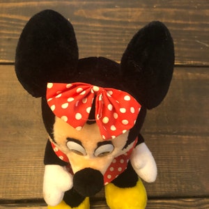 May include: A plush toy of Minnie Mouse with black ears, a red bow with white polka dots, and a red and white polka dot dress. The toy has yellow shoes and a black nose.