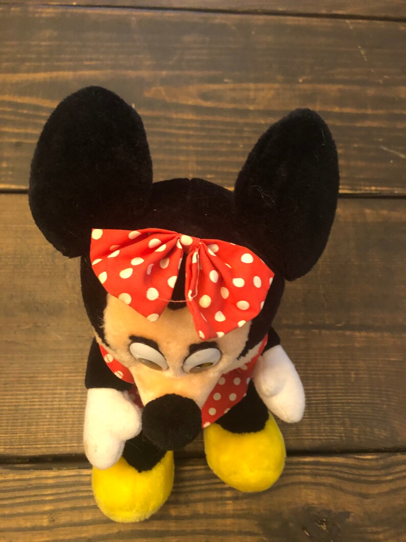 May include: A plush toy of Minnie Mouse with black ears, a red bow with white polka dots, and a red and white polka dot dress. The toy has yellow shoes and a black nose.