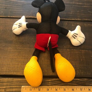 May include: A black and red Mickey Mouse plush toy with a yellow mesh fabric. The toy is about 12 inches tall and has a red and white mesh fabric shorts. The toy is lying on its back with its arms outstretched.