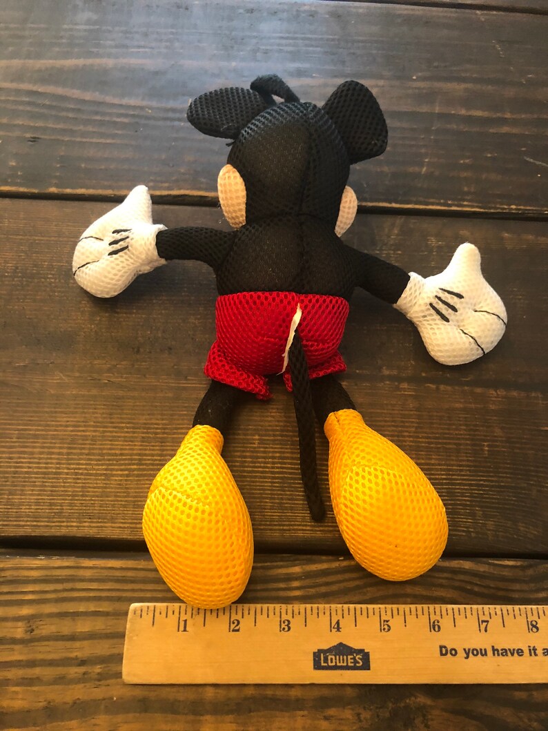 May include: A black and red Mickey Mouse plush toy with a yellow mesh fabric. The toy is about 12 inches tall and has a red and white mesh fabric shorts. The toy is lying on its back with its arms outstretched.