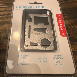 May include: A silver multi-tool with 11 functions, including a bottle opener, a ruler, and a saw. The tool is packaged in a clear plastic case with a white background and the text "SURVIVAL TOOL" and "KIKKERLAND" printed on it.