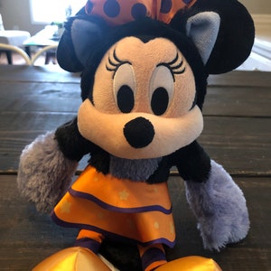 May include: A plush Minnie Mouse doll in a Halloween costume. The doll has a black head and ears, a light-colored face, and a large orange bow with purple polka dots. She wears an orange dress with star accents and gold shoes.