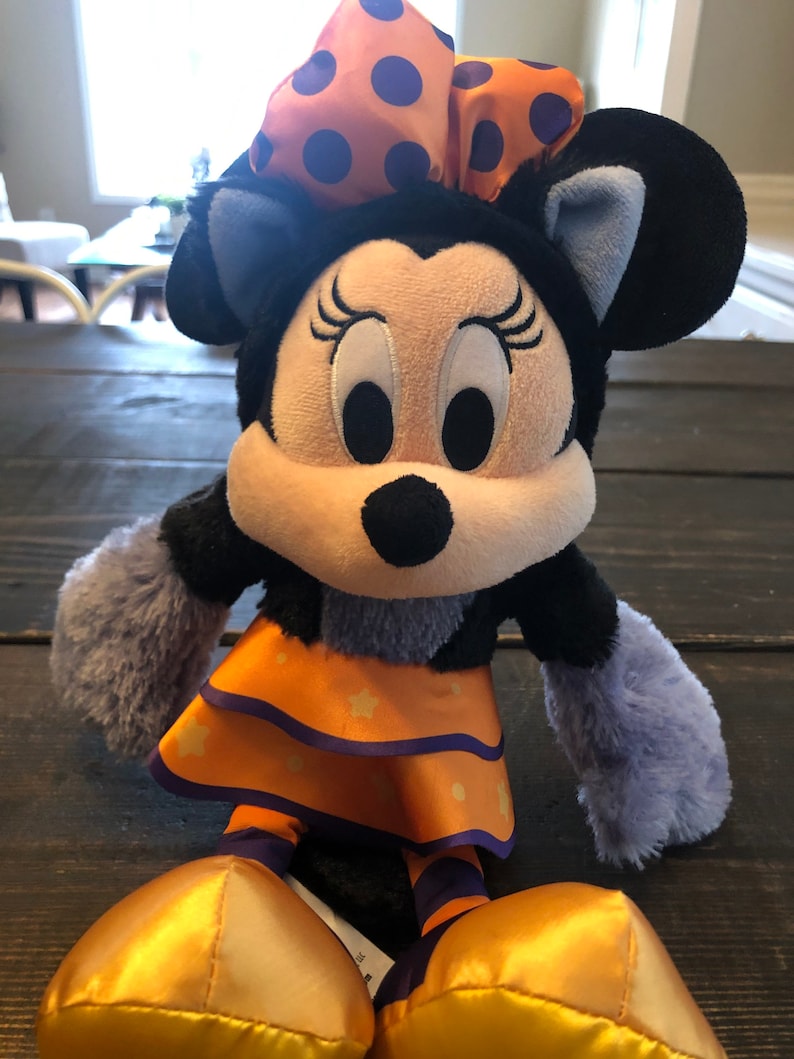 May include: A plush Minnie Mouse doll in a Halloween costume. The doll has a black head and ears, a light-colored face, and a large orange bow with purple polka dots. She wears an orange dress with star accents and gold shoes.