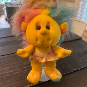 May include: A vintage yellow Troll doll with colorful hair and a polka-dot outfit. The doll has a smiling face, small ears, and is wearing flip-flops. The hair is a mix of pink, yellow, and blue. The doll is standing on a white base.