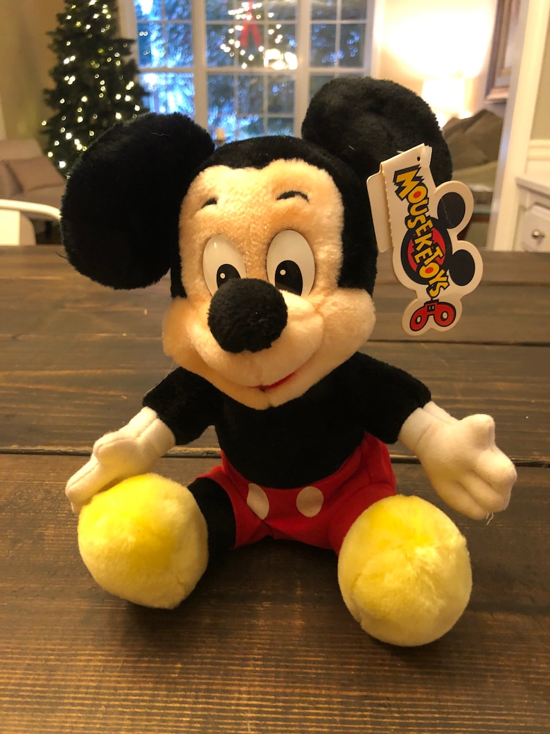 May include: A plush Mickey Mouse toy sitting on a wooden surface. The toy is wearing red shorts with white polka dots and has a yellow tag that says "Mouseketoys".