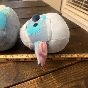 May include: Two plush toys shaped like a cartoon character, one partially visible. The visible toy is light blue with darker blue and teal accents, a dark blue sphere, and large, pink-lined ears. The toy is resting on a dark wooden surface.