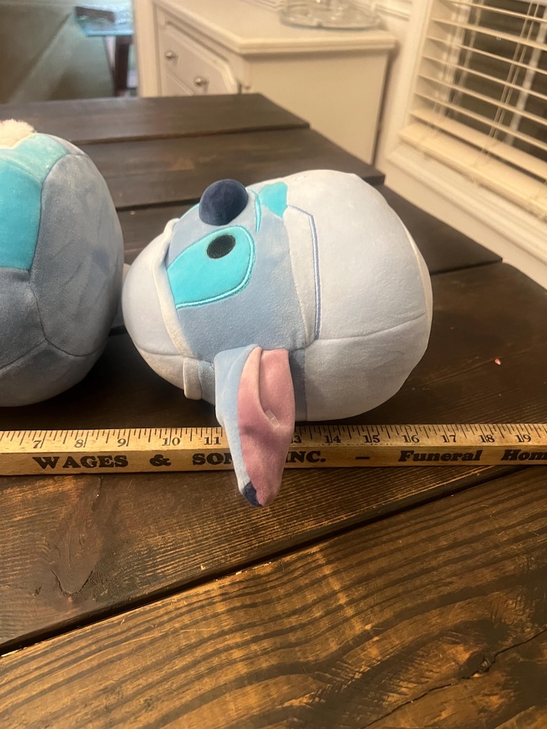 May include: Two plush toys shaped like a cartoon character, one partially visible. The visible toy is light blue with darker blue and teal accents, a dark blue sphere, and large, pink-lined ears. The toy is resting on a dark wooden surface.