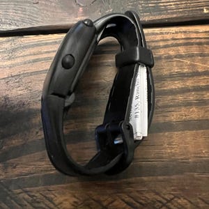 May include: A black fitness tracker with a curved design and a button on the side. The band is made of a flexible material. The text "#135, Rancho Santa" is visible on a small white paper.