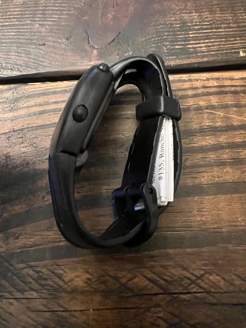 May include: A black fitness tracker with a curved design and a button on the side. The band is made of a flexible material. The text "#135, Rancho Santa" is visible on a small white paper.