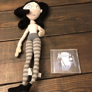 May include: A gray and black plush doll of a character from the movie "Corpse Bride" with striped tights. The doll is lying on a wooden surface next to a CD case for the movie soundtrack.