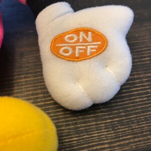 May include: A white plush hand with an orange circle that says "ON OFF" on it. The hand is part of a plush toy.