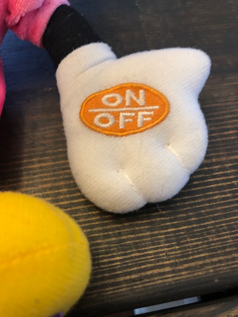 May include: A white plush hand with an orange circle that says "ON OFF" on it. The hand is part of a plush toy.