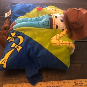 以下が含まれることがあります： トイストーリーのキャラクターが描かれた青、緑、黄色のぬいぐるみ枕。キャラクターは茶色のカウボーイハットをかぶって横になっています。枕の長さは約17インチです。