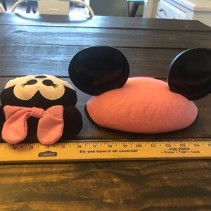 May include: Two plush Minnie Mouse ears, one with a pink headband and black ears, the other with a black headband and pink ears. The ears are on a wooden table with a ruler.