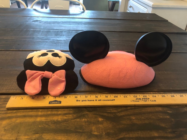 May include: Two plush Minnie Mouse ears, one with a pink headband and black ears, the other with a black headband and pink ears. The ears are on a wooden table with a ruler.