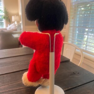 May include: A vintage plush doll with a black head and a red outfit with white trim. The doll is mounted on a white metal stand, showcasing its back and side profile. The doll is on a dark wooden table.