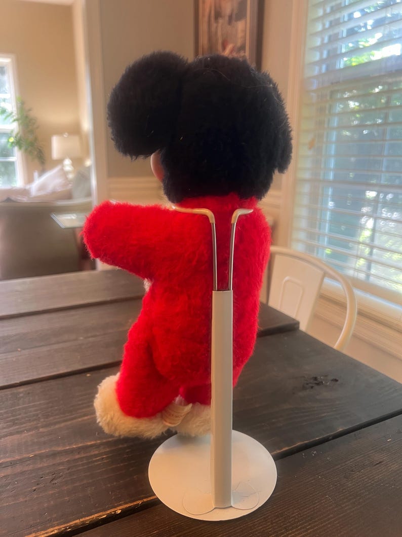 May include: A vintage plush doll with a black head and a red outfit with white trim. The doll is mounted on a white metal stand, showcasing its back and side profile. The doll is on a dark wooden table.