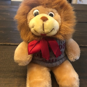 May include: A plush lion toy with a brown mane, wearing a gray sweater with a red bow tie. The lion has large, black eyes and a friendly expression.