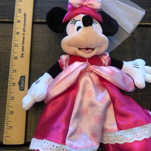 May include: Pink and white plush Minnie Mouse doll wearing a pink dress with white polka dots and a pink crown with a white veil.