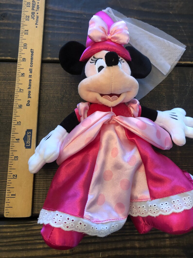 May include: Pink and white plush Minnie Mouse doll wearing a pink dress with white polka dots and a pink crown with a white veil.