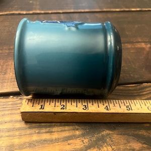 May include: A teal-colored glass jar with a rounded shape. The jar is approximately 5 inches long. A wooden ruler is placed next to the jar for scale. The jar has a glossy finish and appears to be empty.
