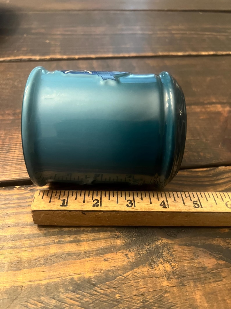 May include: A teal-colored glass jar with a rounded shape. The jar is approximately 5 inches long. A wooden ruler is placed next to the jar for scale. The jar has a glossy finish and appears to be empty.