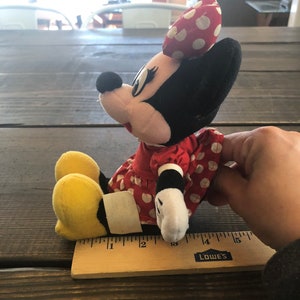 Puede incluir: Un vestido rojo con lunares blancos y un lazo amarillo en la cabeza. El peluche est&aacute; sentado sobre una superficie de madera con una regla a su lado. La regla est&aacute; marcada en pulgadas y tiene la palabra "LOWE'S" impresa en ella.