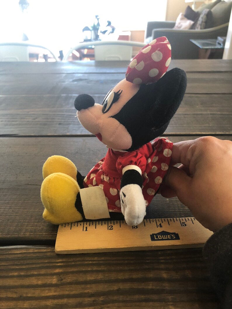 Puede incluir: Un vestido rojo con lunares blancos y un lazo amarillo en la cabeza. El peluche est&aacute; sentado sobre una superficie de madera con una regla a su lado. La regla est&aacute; marcada en pulgadas y tiene la palabra "LOWE'S" impresa en ella.