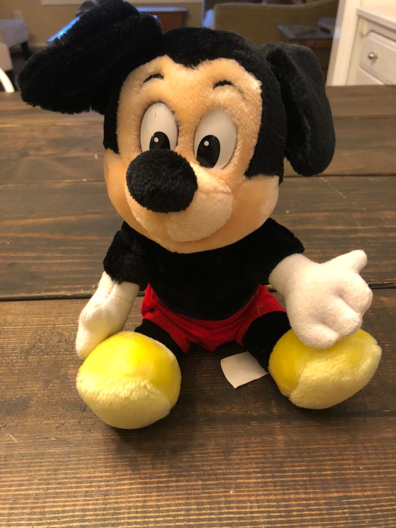 Puede incluir: Un juguete de peluche de Mickey Mouse con orejas negras, nariz negra y zapatos amarillos. El juguete lleva pantalones cortos rojos y tiene una barriga blanca.