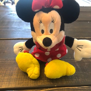 May include: A plush toy of Minnie Mouse wearing a red dress with white polka dots and a red bow with white polka dots. The toy has black ears and yellow feet.