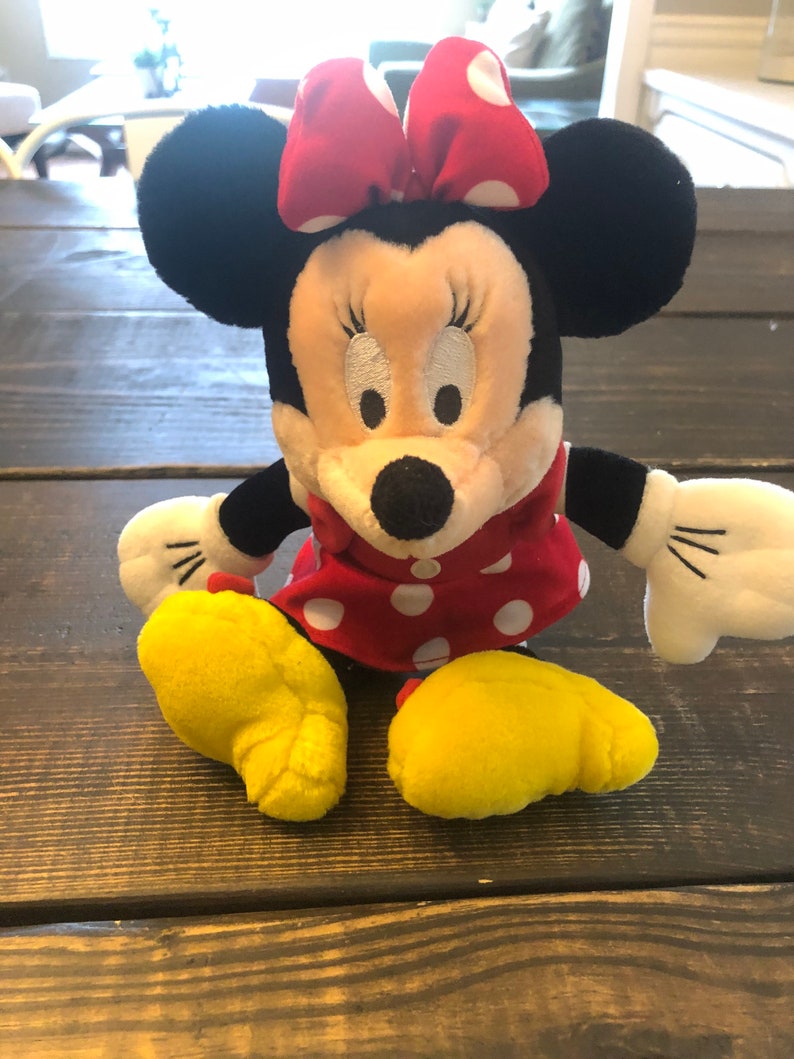 May include: A plush toy of Minnie Mouse wearing a red dress with white polka dots and a red bow with white polka dots. The toy has black ears and yellow feet.