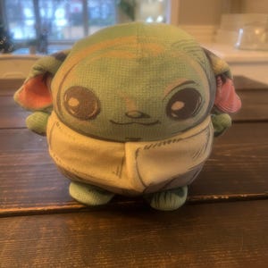 May include: A plush toy of a character with large eyes, green skin, and large ears. The toy is wearing a tan-colored outfit and has a smiling expression. The toy is sitting on a dark wooden surface.