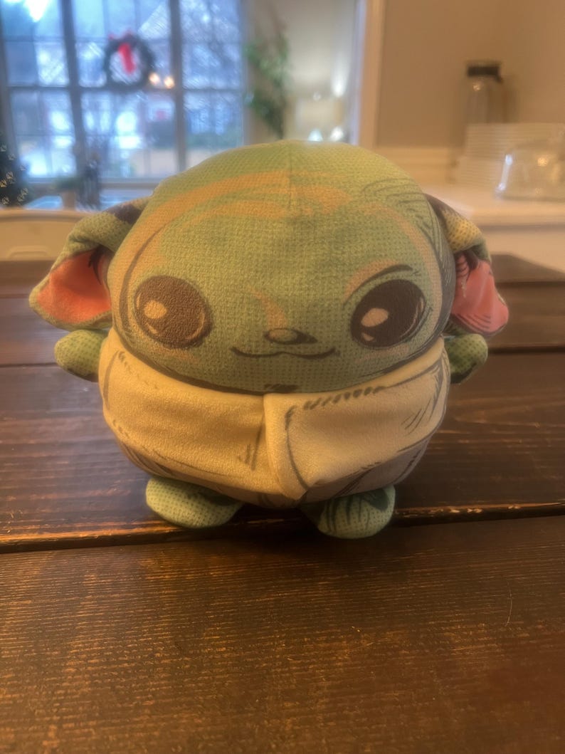 Pode incluir: Um brinquedo de pel&uacute;cia verde de um personagem com olhos e orelhas grandes, vestindo uma roupa bege. O brinquedo tem uma express&atilde;o sorridente e est&aacute; posicionado em uma superf&iacute;cie de madeira escura. Personagem popular.
