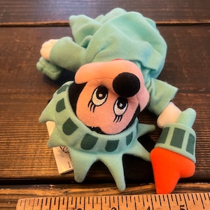 May include: A plush toy of Mickey Mouse dressed as the Statue of Liberty. The toy is green, orange, and black. The toy is about 5 inches tall.
