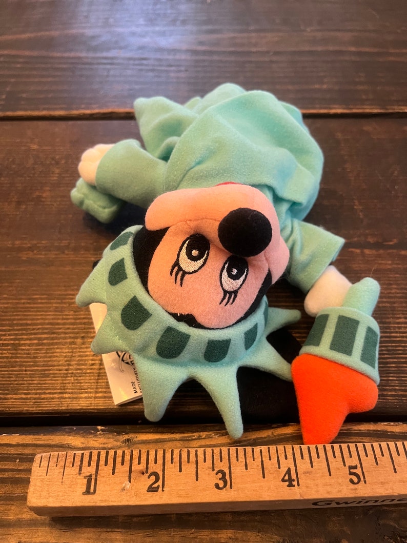 May include: A plush toy of Mickey Mouse dressed as the Statue of Liberty. The toy is green, orange, and black. The toy is about 5 inches tall.
