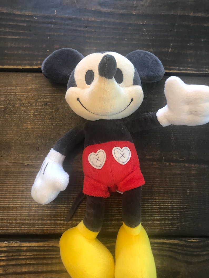 Puede incluir: Un juguete de peluche de Mickey Mouse con pelaje negro, guantes blancos, zapatos amarillos y pantalones cortos rojos con corazones blancos. El juguete tiene una sonrisa cosida y una nariz negra.