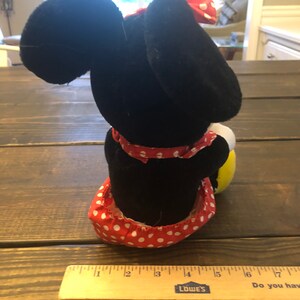 May include: A black and red plush toy of a cartoon mouse wearing a red and white polka dot dress and a red bow. The toy is sitting on a wooden surface.