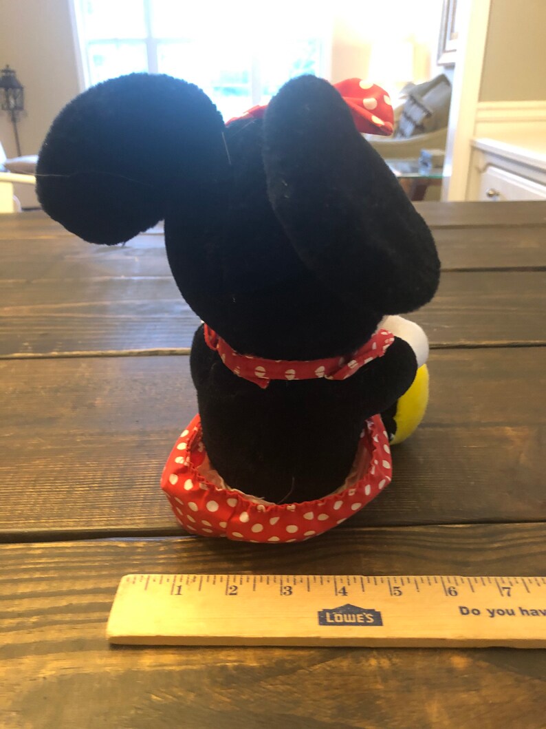 May include: A black and red plush toy of a cartoon mouse wearing a red and white polka dot dress and a red bow. The toy is sitting on a wooden surface.